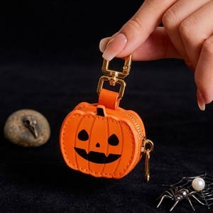 May include: A bright orange pumpkin-shaped pouch with a black jack-o'-lantern face, featuring a gold-toned metal lobster clasp and a side zipper closure. The small accessory is constructed with stitched edges and a matching orange leather-like material.
