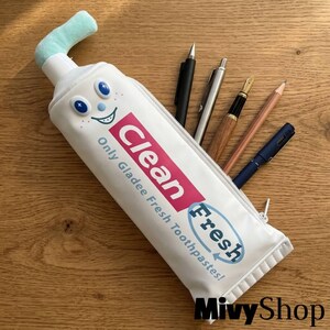 May include: A white pencil case shaped like a toothpaste tube with a light blue plush top. The front features a cartoon face and the text Clean Fresh Only Gladee Fresh Toothpastes! Five various pens and pencils are tucked inside the open top.