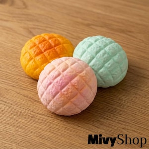 May include: Three round, textured squishy toys resembling melon pan bread. One is bright orange, one is pale mint green, and one is a soft gradient of pink and orange. Each features a distinct grid pattern across the rounded surface.
