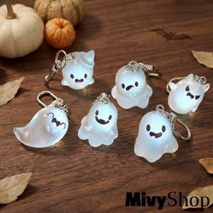 May include: Six translucent white ghost keychains with various facial expressions, each featuring a silver-toned metal lobster clasp. The ghosts have smooth, rounded forms and are illuminated, displaying distinct eyes and mouths on their surfaces.