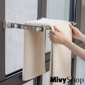 Puede incluir: Una barra de tensión de metal plateado montada entre marcos de ventana, con una serie de recortes circulares en el lateral. Una toalla beige con textura de gofre está colgada sobre la barra, asegurada por tapas de plástico blanco.