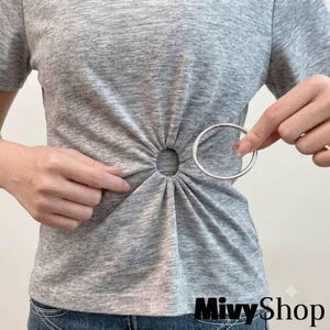 May include: A heather gray short-sleeved t-shirt featuring a central circular cutout detail at the waist. The fabric is gathered around a smooth, silver-toned metal ring that holds the material in place, creating a sunburst effect of pleats.