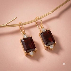 Puede incluir: Un par de pendientes colgantes de oro con piedras preciosas de granate rojo oscuro rectangulares. Cada pendiente está adornado con dos pequeñas piedras redondas transparentes en la parte superior e inferior, suspendidas de un gancho de oro.