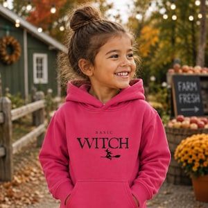 May include: A bright pink hooded sweatshirt featuring the text "BASIC WITCH" in black lettering, accompanied by a small black graphic of a witch flying on a broomstick. The garment includes a front kangaroo pocket and long sleeves.
