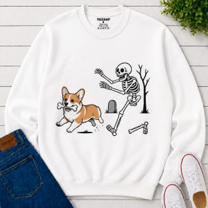 May include: A white long-sleeve sweatshirt featuring a graphic print of a brown and white Corgi dog running with a bone in its mouth, followed by a black and white skeleton reaching out, with a tombstone and a bare tree in the background.