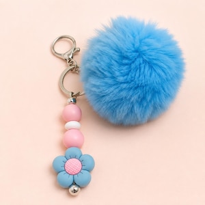 May include: A keychain featuring a large, fluffy, bright blue pom-pom attached to a silver-toned metal ring. Below the ring, a vertical strand consists of two round pink beads, one small white bead, and a blue flower charm with a pink center.