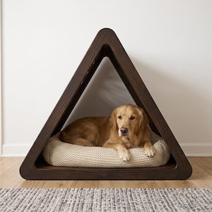 May include: A dark brown wooden pet house with a triangular frame. Inside the structure, a golden-colored dog with long fur is lying down on a thick, cream-colored, textured knit cushion that fills the base of the triangular enclosure.