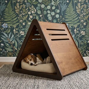 May include: A triangular wooden pet house with a smooth brown finish and three horizontal ventilation slats on the side. Inside the structure, a beagle dog with brown, black, and white fur is sleeping on a textured beige rectangular cushion.