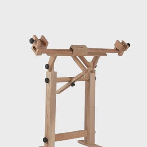 May include: A light-colored wooden floor stand featuring an adjustable horizontal frame with two side clamps. The structure includes vertical supports, a central angled brace, and black adjustment knobs for securing the frame at various positions.