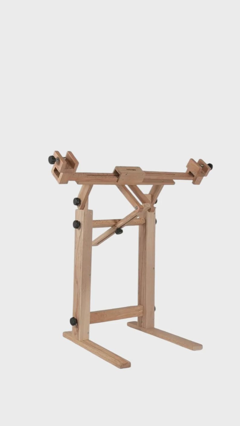 May include: A light-colored wooden floor stand featuring an adjustable horizontal frame with two side clamps. The structure includes vertical supports, a central angled brace, and black adjustment knobs for securing the frame at various positions.