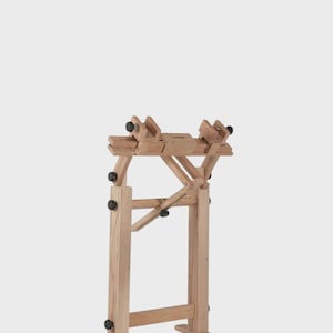 May include: A light-colored wooden craft stand featuring a vertical frame with two horizontal base supports. The top section includes adjustable clamps with black knobs, allowing for positioning and securing items during various manual tasks.