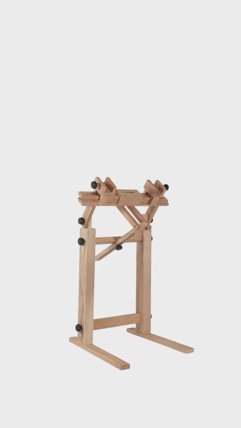 May include: A light-colored wooden craft stand featuring a vertical frame with two horizontal base supports. The top section includes adjustable clamps with black knobs, allowing for positioning and securing items during various manual tasks.