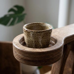 May include: A small, speckled beige ceramic cup sits inside a circular cutout on a wooden armrest. The cup has a textured, earthy appearance with dark brown flecks, while the armrest features visible layers of wood grain in a medium brown tone.