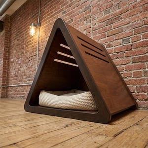 May include: A triangular pet house constructed from dark brown wood with a smooth finish. The structure features three horizontal ventilation slats on the side panel and contains a soft, cream-colored, textured knit cushion inside the base.