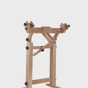 May include: A light-colored wooden floor stand featuring an adjustable frame with multiple black knobs for securing items. The structure includes two vertical supports, a horizontal top bar with sliding components, and two horizontal base feet.