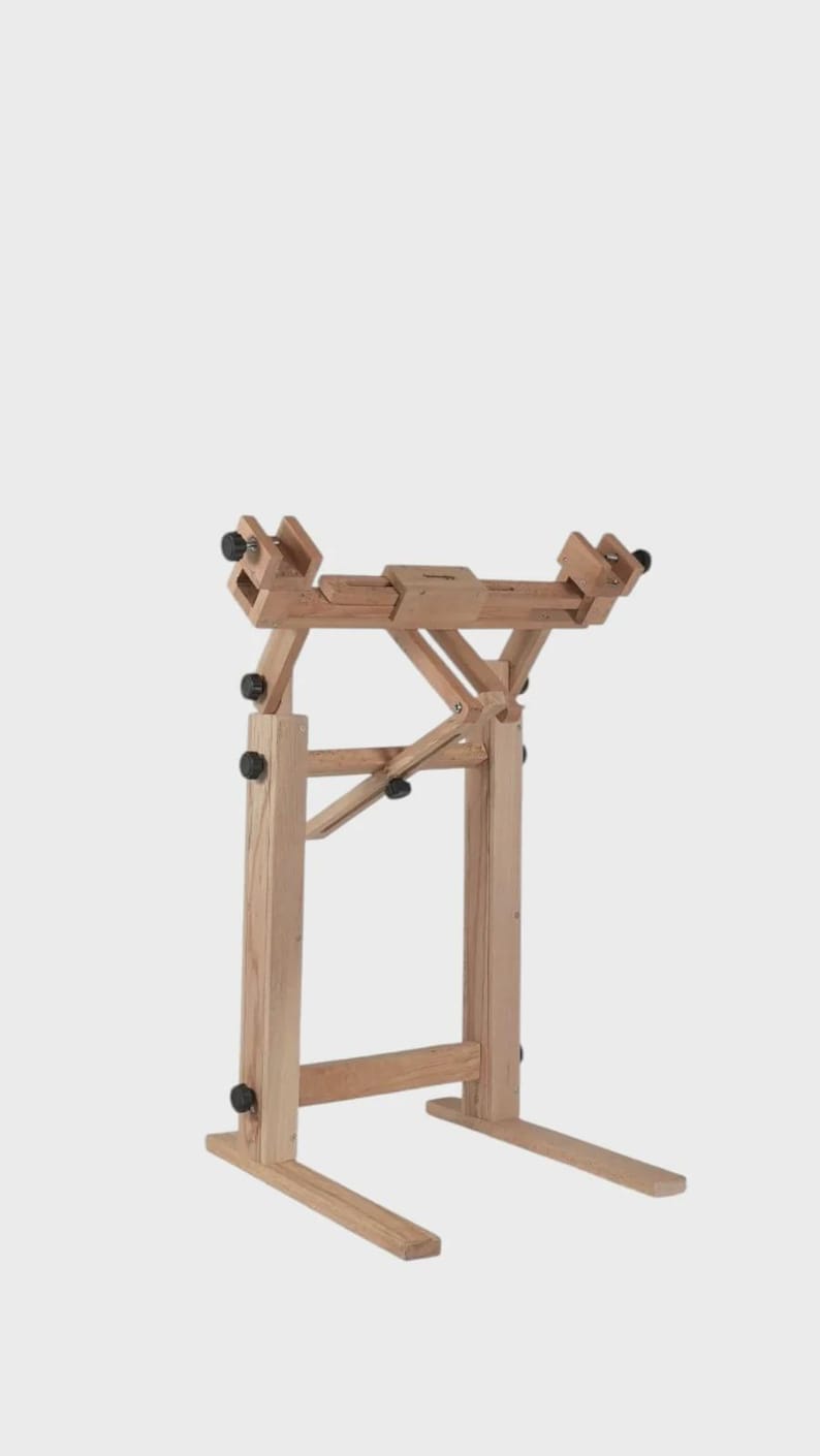 May include: A light-colored wooden floor stand featuring an adjustable frame with multiple black knobs for securing items. The structure includes two vertical supports, a horizontal top bar with sliding components, and two horizontal base feet.