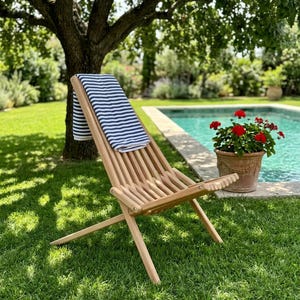May include: A light-colored wooden folding chair with a slatted seat and backrest, draped with a white towel featuring horizontal navy blue stripes. The chair is positioned on a green lawn, showcasing its simple, functional design and structure.
