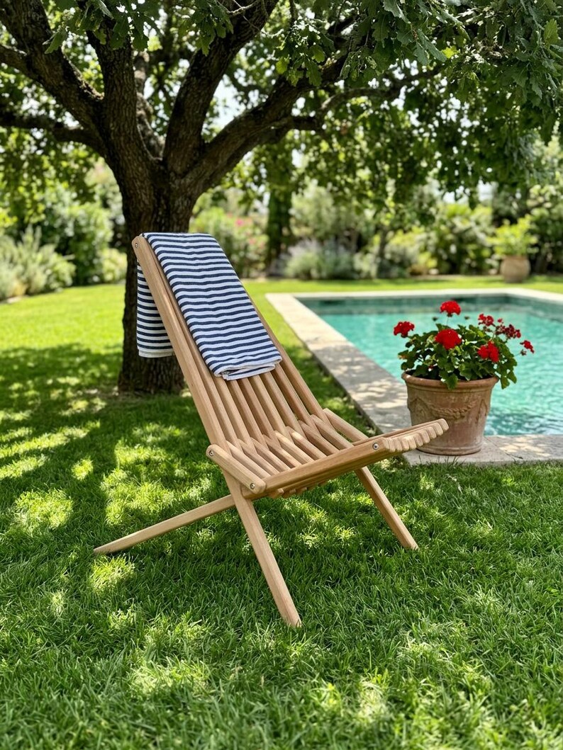 May include: A light-colored wooden folding chair with a slatted seat and backrest, draped with a white towel featuring horizontal navy blue stripes. The chair is positioned on a green lawn, showcasing its simple, functional design and structure.