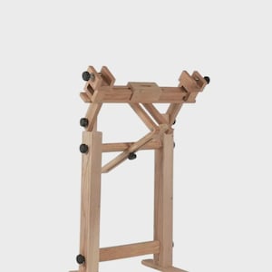 May include: A light-colored wooden floor stand featuring an adjustable frame with multiple black knobs for securing items. The structure consists of two vertical supports, a horizontal crossbar, and a base with two extending feet for stability.