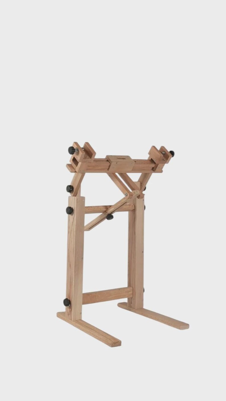 May include: A light-colored wooden floor stand featuring an adjustable frame with multiple black knobs for securing items. The structure consists of two vertical supports, a horizontal crossbar, and a base with two extending feet for stability.