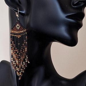 May include: A long, triangular beaded earring featuring a geometric pattern in black, bronze, and gold tones. The earring is constructed from small seed beads and longer tube beads, creating a layered fringe effect that hangs from a metal hook.