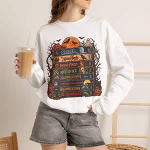 May include: A white long-sleeve sweatshirt featuring a graphic print of a stack of VHS tapes with titles including Casper, The Addams Family, Hocus Pocus, Beetlejuice, Halloweentown II, The Nightmare Before Christmas, Halloweentown, and Goosebumps.