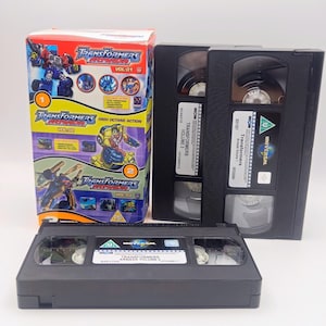 May include: A Transformers Armada VHS collection featuring a cardboard box set with colorful character graphics and text, alongside three black VHS tapes labeled Volume 1, Volume 2, and Volume 3, each with Universal branding and clear film reels.
