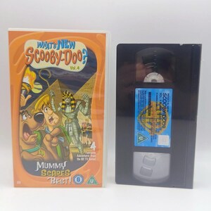 May include: A What's New Scooby-Doo? Vol. 4 Mummy Scares Best! VHS tape and its orange plastic case. The cover features Scooby-Doo, Shaggy, and a mummy in front of pyramids. The black VHS tape has a blue label with text and a green U rating symbol.