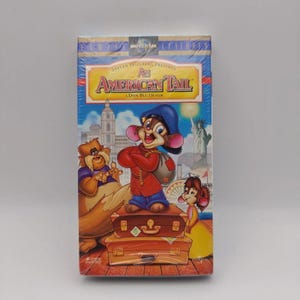 Pode incluir: Uma fita VHS selada do filme de animação An American Tail, apresentando um rato de desenho animado com chapéu azul e casaco vermelho em cima de duas malas castanhas empilhadas, com um gato grande e um rato mais pequeno num cenário urbano.