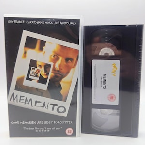 May include: A Memento VHS tape in a plastic case. The cover features the title Memento, the text Some memories are best forgotten, and a photo of Guy Pearce and Carrie-Anne Moss. The black VHS cassette has a white label with the text Pathe, Memento, and VFC23189.