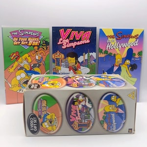 May include: A 3-video gift set featuring The Simpsons. The box displays three oval windows showing artwork from the included VHS tapes: On Your Marks, Get Set, D'oh!, Viva Los Simpsons, and Go to Hollywood. The three individual VHS cases stand behind the box.