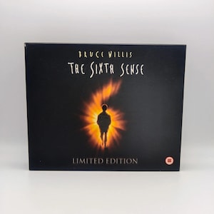 May include: A black rectangular DVD case for the film The Sixth Sense, featuring the text Bruce Willis and The Sixth Sense in white lettering. A silhouette of a person stands in the centre surrounded by a bright orange and yellow light.