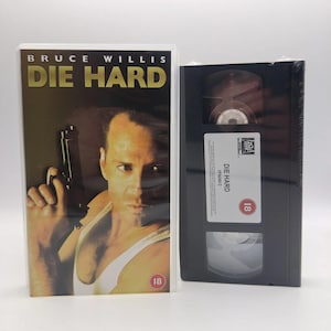 May include: A Die Hard VHS tape in a clear plastic case next to a black VHS cassette. The cover features Bruce Willis holding a handgun, wearing a white tank top, with the title Die Hard in gold text. The cassette has a white label with text.