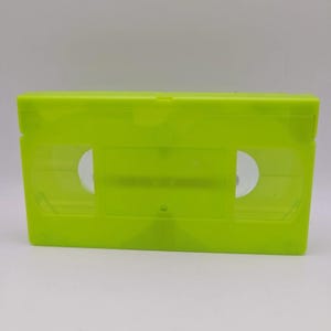 Könnte beinhalten: Eine leuchtend neongrüne VHS-Kassette mit einem durchscheinenden Kunststoffgehäuse. Das rechteckige Speichermedium verfügt über zwei kreisförmige Spulen, die durch das klare Sichtfenster auf der Vorderseite erkennbar sind und ein einfarbiges Design aufweisen.
