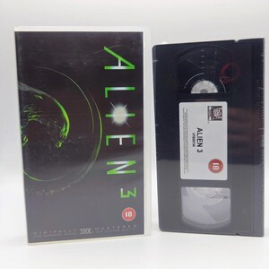 May include: A VHS tape and its plastic case for the film Alien 3. The black case features the title in green text, a graphic of an alien, and an 18 age rating. The black VHS tape has a white label with the text Alien 3, an 18 rating, and 20th Century Fox logo.