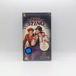 May include: A VHS tape of the film The Sting, 25th Anniversary Edition. The cover features an illustration of two men in 1930s attire, with the title in red text, a PG rating, and a blue Retro Movies Verse sticker on the lower left corner.