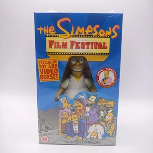 May include: A blue box set titled The Simpsons Film Festival featuring a Homer Simpson figure visible through a cutout. The packaging includes text stating Exclusive Toy and Video Boxset and Special Featurette with Troy McClure, plus illustrations of various characters.
