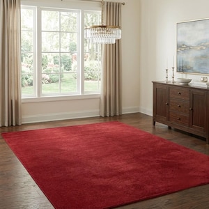 May include: A large, rectangular area rug in a solid, deep red color with a short, dense pile texture. The rug is positioned flat on a dark brown hardwood floor, covering a significant portion of the floor space in a room with neutral walls.