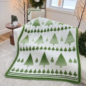 May include: A white crochet blanket featuring a repeating pattern of green pine trees in varying sizes. The blanket has a solid green border and horizontal stripes along the bottom edge, crafted with a textured yarn finish throughout the design.