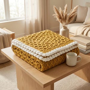 May include: A folded mustard yellow crochet blanket with a textured honeycomb pattern and a thick, white, bobble-stitch border. The blanket is square-shaped and features a consistent, repeating geometric design throughout the fabric.