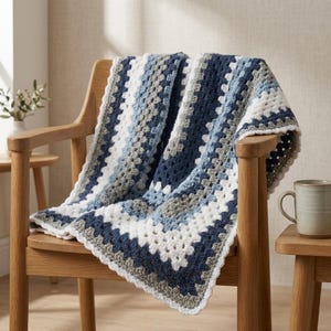 Puede incluir: Una manta tejida a mano con un patrón clásico de cuadrados de la abuela en una paleta de colores azul marino, azul claro, gris y blanco. La manta está colocada sobre una silla de madera, mostrando su textura de hilo y sus bordes festoneados.