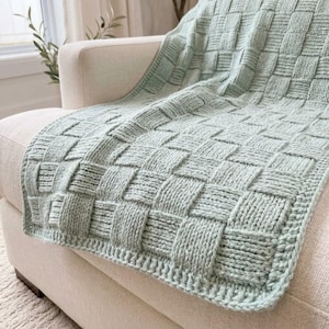 May include: A light sage green knitted throw blanket featuring a textured basketweave pattern. The blanket is constructed with thick yarn, creating a repeating square grid design that covers the entire surface of the soft textile piece.