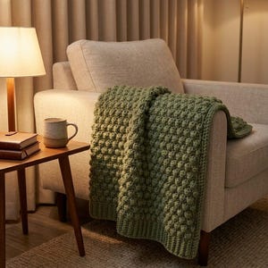 Può includere: Una coperta all'uncinetto color verde salvia caratterizzata da una texture a rilievo con punti a nocciolina su tutta la superficie e un bordo a coste sul fondo. La coperta è drappeggiata sulla seduta e sul bracciolo di una poltrona beige.