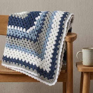 Può includere: Una coperta lavorata all'uncinetto a mano con un classico motivo a piastrelle granny in una palette di colori blu navy, azzurro, grigio e bianco. La coperta è realizzata con file ripetute di punti e rifinita con un bordo bianco smerlato.