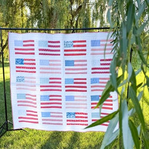 May include: A rectangular quilt featuring a repeating pattern of twenty American flag motifs arranged in five rows and four columns. Each flag block is constructed from various red, white, and blue patterned fabrics with visible stitching.