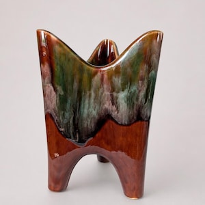 May include: A ceramic vase featuring a glossy finish with a deep brown base that transitions into a mottled green and brown glaze at the top. The vessel has a distinct four-legged base and a scalloped, undulating rim with four upward-pointing peaks.