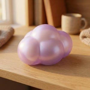 May include: A smooth, rounded, cloud-shaped object with a pearlescent, light purple finish. The surface has a soft, reflective sheen, and the form consists of multiple interconnected, bulbous segments that create a stylized, puffy appearance.