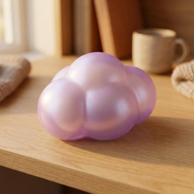 May include: A smooth, rounded, cloud-shaped object with a pearlescent, light purple finish. The surface has a soft, reflective sheen, and the form consists of multiple interconnected, bulbous segments that create a stylized, puffy appearance.