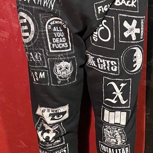 May include: Black sweatpants adorned with numerous white patches displaying diverse graphics and text. Patches include the words "DISTAWN", "MEMORY OF ALL YOU DEAD FUCKS", and "TOTALITAR". The sweatpants have a relaxed fit.