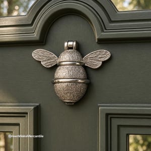 May include: A silver-toned bee-shaped door knocker with a textured body and wings, mounted on a dark olive green paneled door. The metal insect features a hinged top section for knocking and is centered on the door surface. Text reads CrownWakeMercantile.